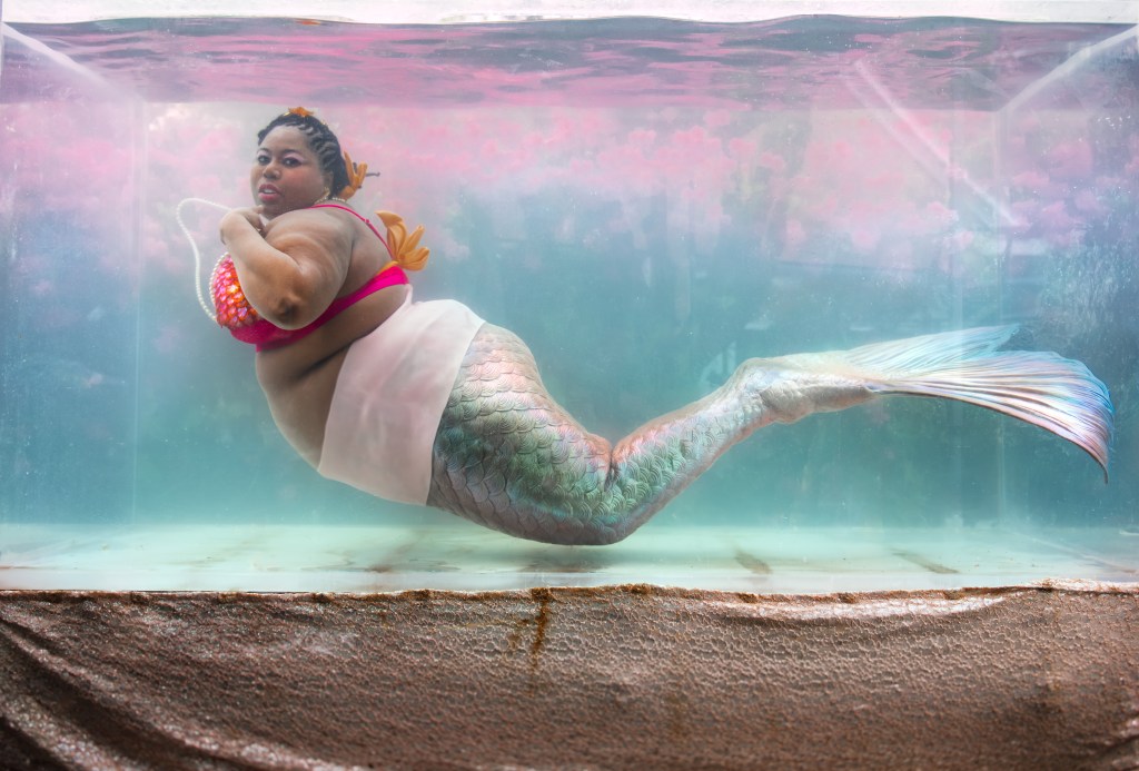 Mermaid Chè is in a tank. Her hair is cornrowed with an orange starfish clip. White Pearls float above her chest. She is looks at you with a relaxed slightly sultry expression. Her top has shimmering orange and pink scales layered over a bright fuchsia base. There is a light pink band at her large waist. Her tail is iridescent with hints of green, pink, blue and brown. 